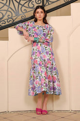 A woman wearing a white dress with a soft lilac floral print, featuring a square neckline, elastic detailing at the waist, and flared bottom hem. She is also wearing pink heels.